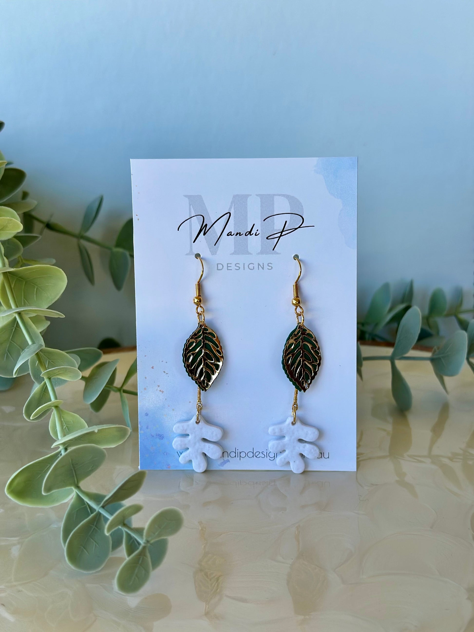 Handmade polymer clay dangle earrings with white RicRac leaf design, gold filigree leaf, and green metal accent – displayed on Mandi P Designs card