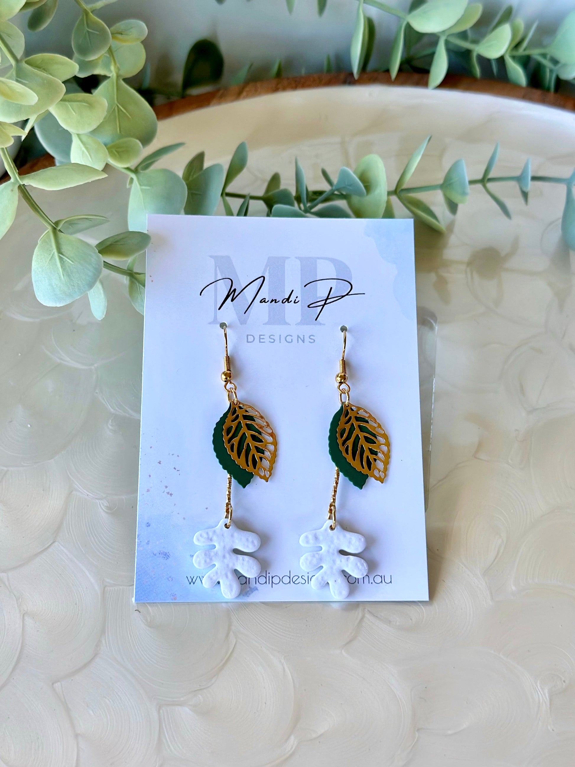 Handmade polymer clay dangle earrings with white RicRac leaf design, gold filigree leaf, and green metal accent – displayed on Mandi P Designs card