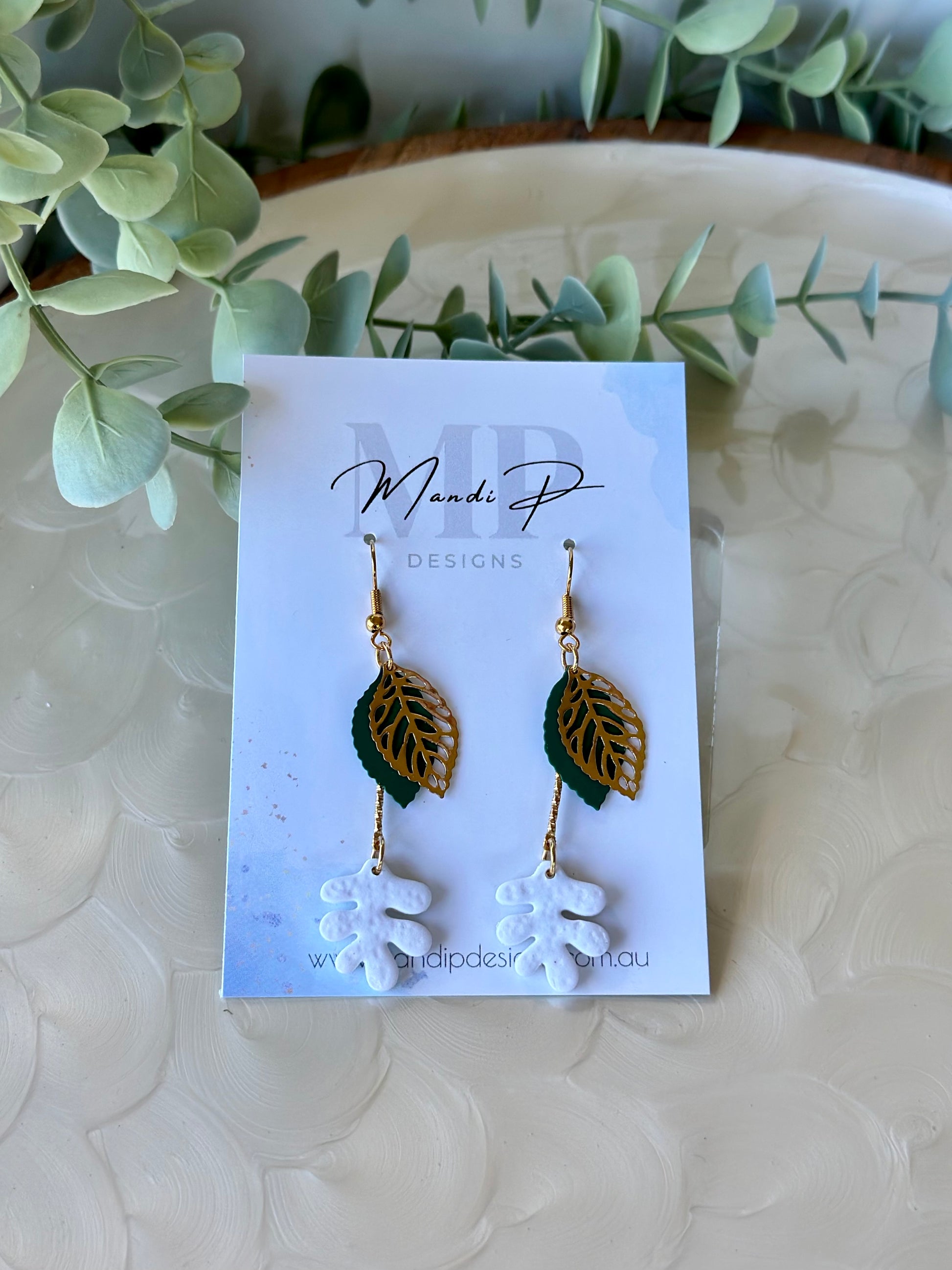 Handmade polymer clay dangle earrings with white RicRac leaf design, gold filigree leaf, and green metal accent – displayed on Mandi P Designs card