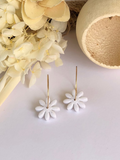 A pair of chalk-colored earrings with gold plated stainless steel hoops and a floral daisy charm, displayed next to a decorative flower arrangement and a hollow sphere.