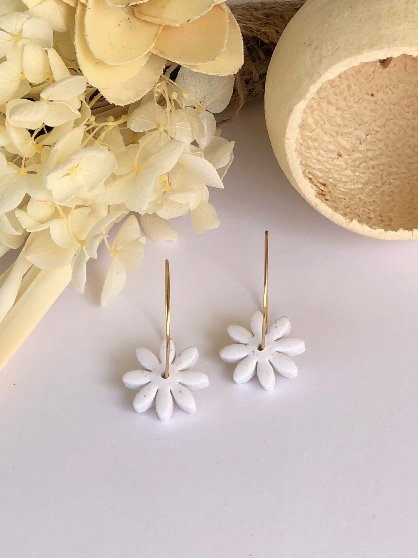 A pair of chalk-colored earrings with gold plated stainless steel hoops and a floral daisy charm, displayed next to a decorative flower arrangement and a hollow sphere.